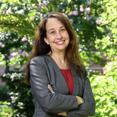 woman with long brown hair smiling at the camera with arms crossed. Wearing a red blouse and black blazer with small white check pattern standing outdoors against a backdrop of green foliage