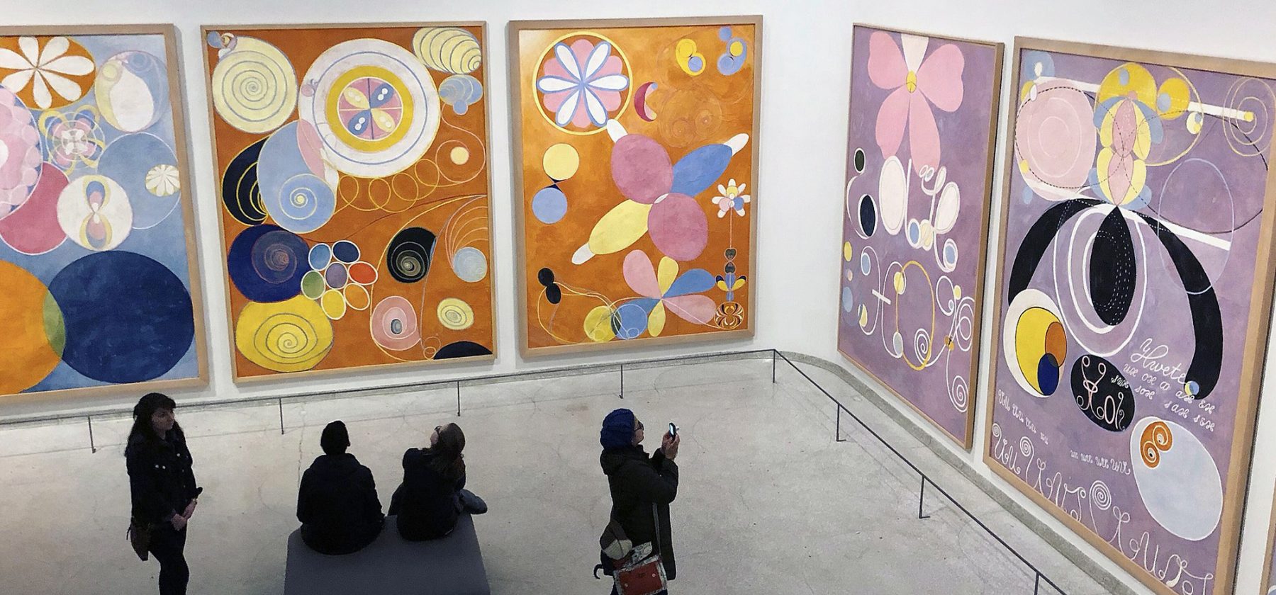 This is a photo of a woman taking a picture on her phone of Hilma af Klint’s “The Ten Largest” at the Solomon R. Guggenheim Museum in New York