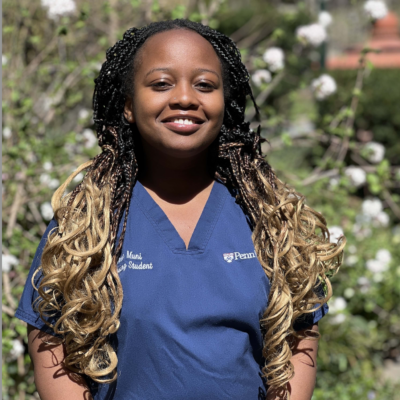 Woman with long hair smiling at the camera wearing a blue shirt that says Penn Nursing. Photos is taken outdoors against greenery with white flowers.