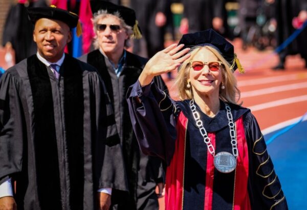 This is a photo of Amy Gutmann, waving to the crowd in graduation regalia. Vice Provost Wendell Pritchett is standing behind her.