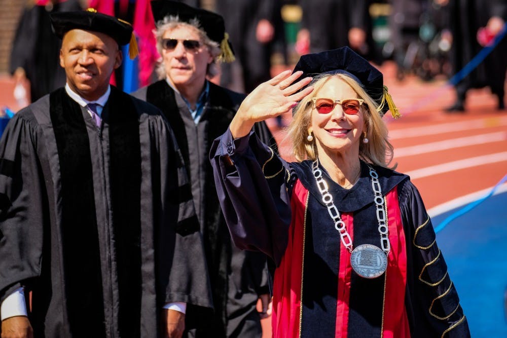 This is a photo of Amy Gutmann, waving to the crowd in graduation regalia. Vice Provost Wendell Pritchett is standing behind her.