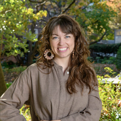 woman with long brown hair and light skin smiling at the camera standing outdoors near a tree, wearing a light brown long-sleeved top