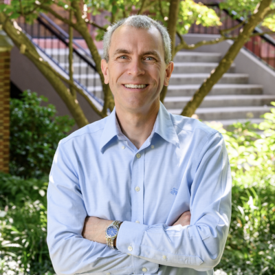 Man with short grey hair smiling at the camera wearing a light blue button down long-sleeve shirt with arms crossed. They are standing against the backdrop of a campus building and green foliage.