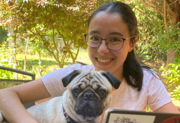 This is a picture of Catherine Michelutti, holding her dog which is a Pug breed, smiling at camera with greenery in the background.