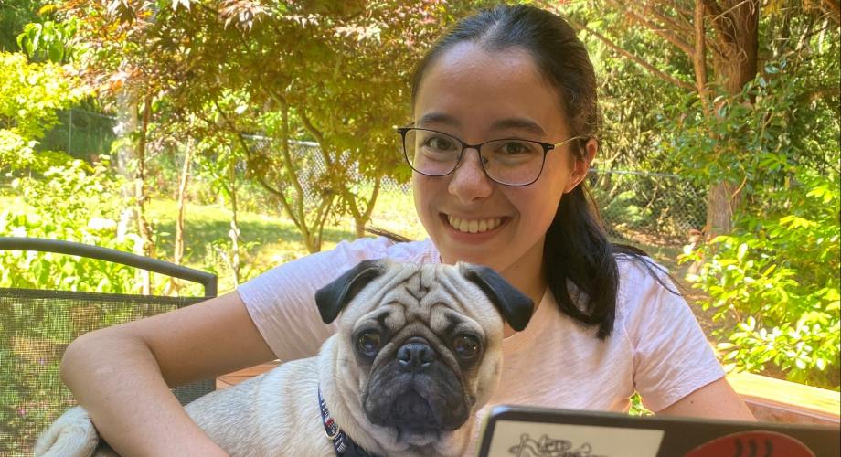 This is a picture of Catherine Michelutti, holding her dog which is a Pug breed, smiling at camera with greenery in the background.