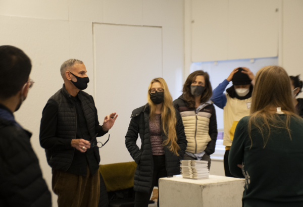 Ernesto Pujol is standing in the middle of a room, surrounded by 6 people looking at him as he is speaking. Everyone in the room has a mask on and Pujol has his hands raised at his waist.