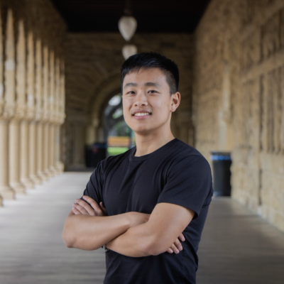 male of Asian background with short black hair wearing black short-sleeved shirt smiling at camera with arms folded standing against a backdrop of an outdoor campus walkway with columns on one side and stone wall on other.