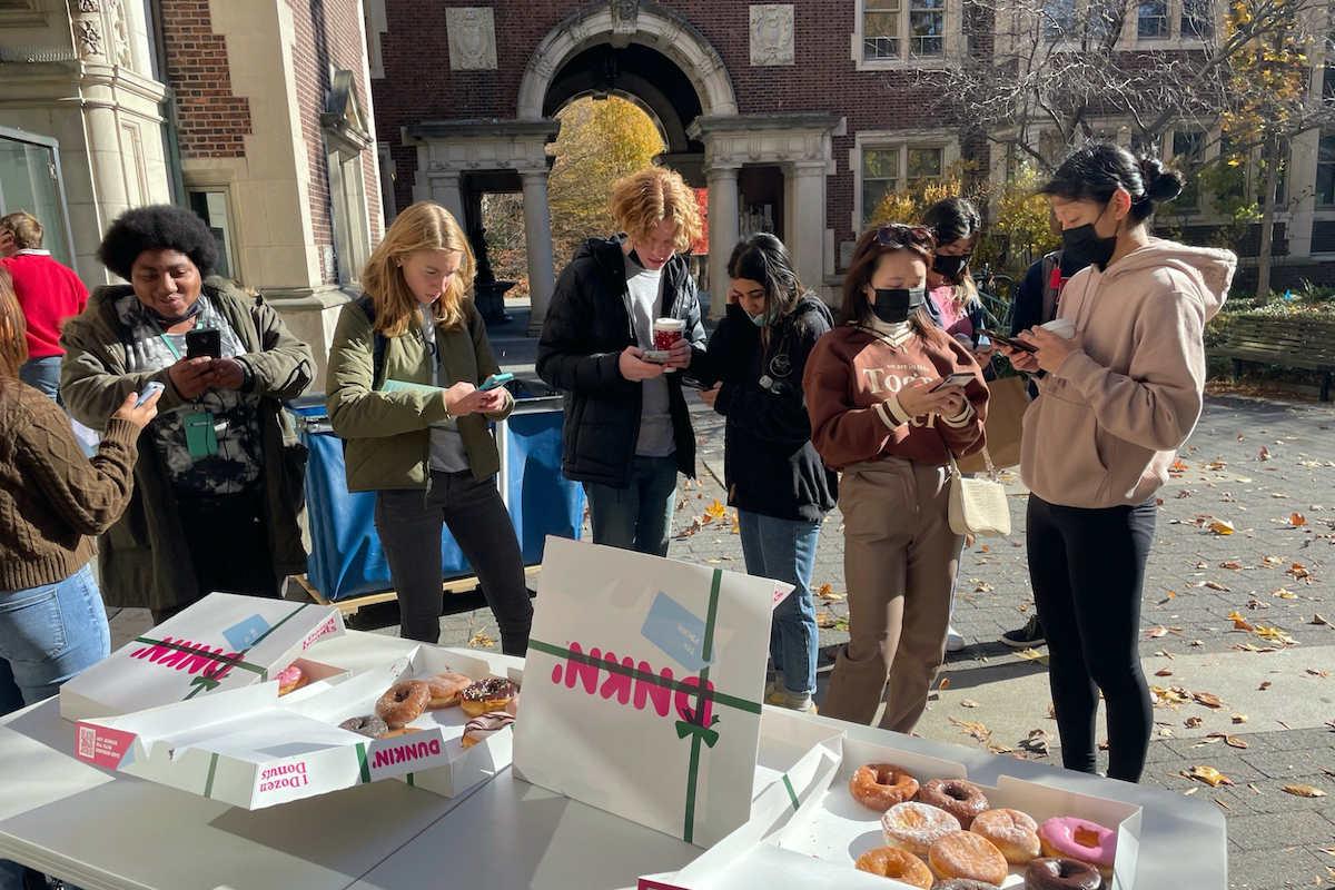 This photo has seven individuals all on their phones outside. They are standing alongside a table with a Dunkin Donuts box opens and donuts on the table.