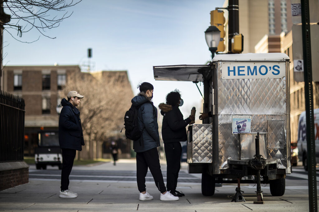 There is a metal food truck with Hemo's on the side and three students in line in front of it. 