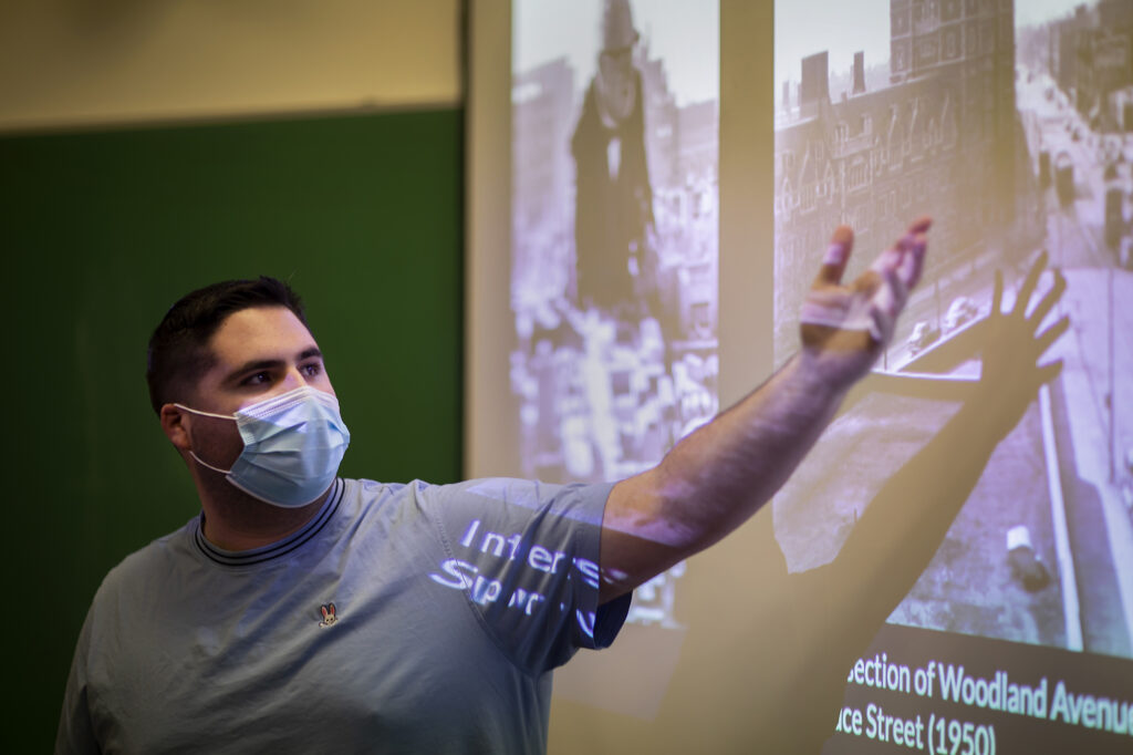 There is a student standing in from of a screen with his arm raised towards the image on the screen. 