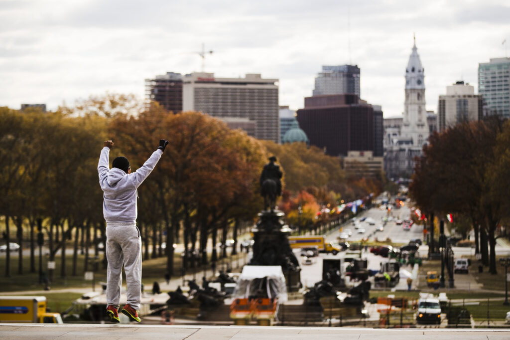 There is a person with their arms up and one black leather glove on with their arms raised as they overlook the city from the top of the Rocky Steps.