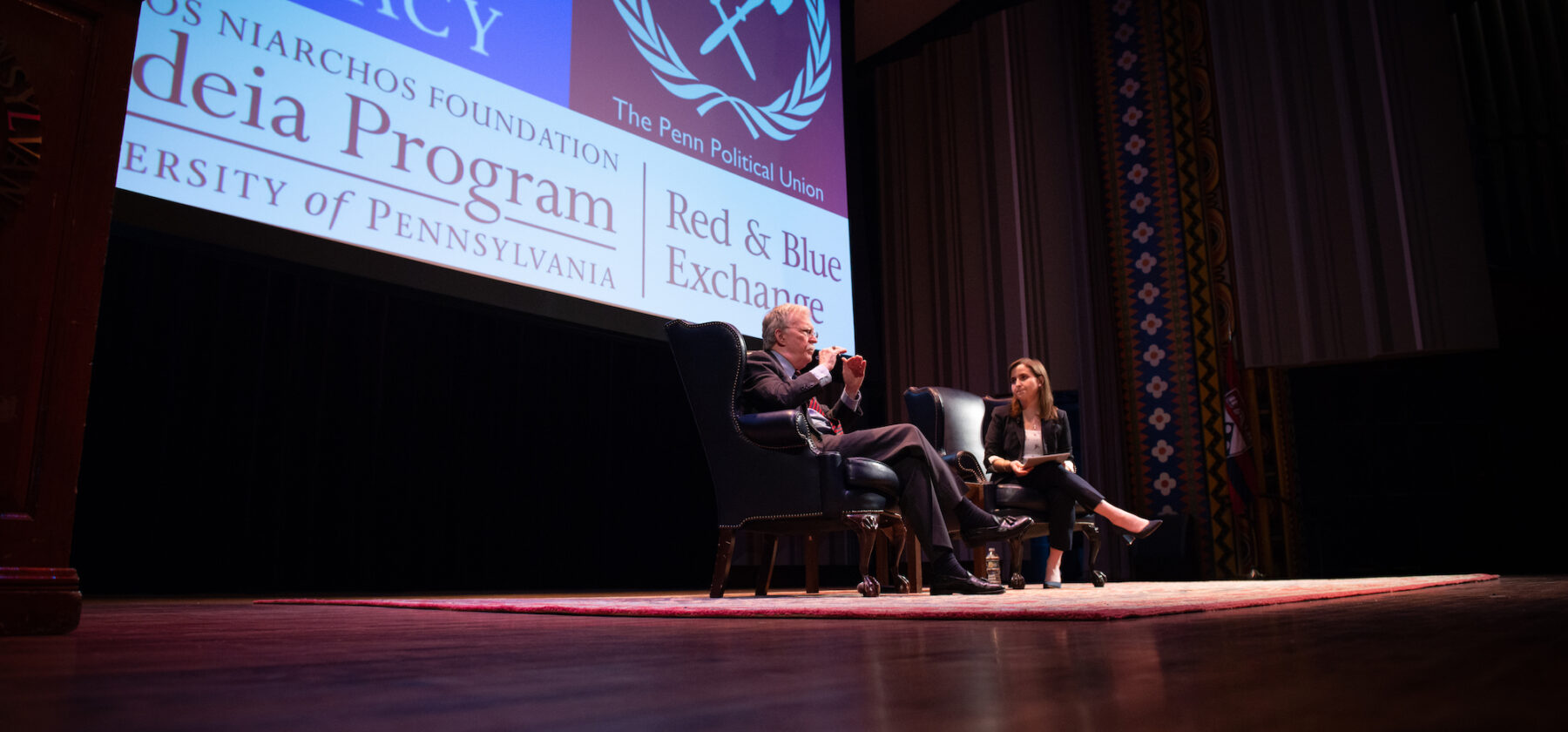 John Bolton sitting next to Lexi Boccuzzi on a stage.