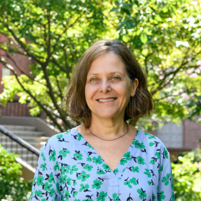 woman with short light brown hair smiling at the camera wearing a light blue, green and black floral blouse. Standing against a backdrop of green foliage and a campus building.