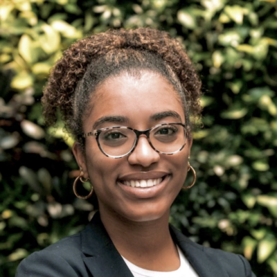 female student with dark skin and light brown hair pulled back wearing glasses, a white shirt and black blazer smiling for the camera