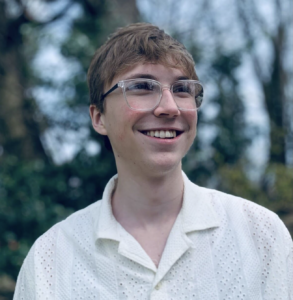 Headshot of person with short blonish brown hair wearing glasses and smiling while looking away from camera. They are standing outdoors again trees and greenery.