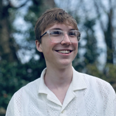 Headshot of person with short blonish brown hair wearing glasses and smiling while looking away from camera. They are standing outdoors again trees and greenery.