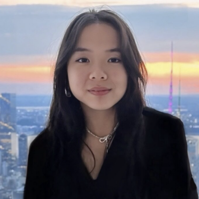 Headshot of woman with long black hair smiling at the camera wearing a black blazer against a window with the sun setting