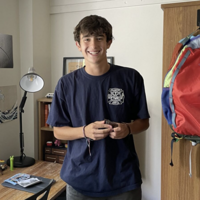Man with short dark brown hair standing in dorm room smiling at camera wearing navy short sleeved shirt