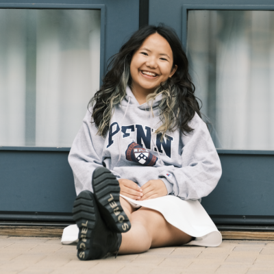 Woman with long black hair seated outdoors on ground smiling at camera wearing a Penn sweatshirt