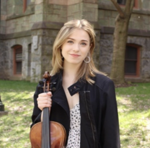 Woman standing outdoors with long blondish brown hair smiling at camera holding a violin in her right hand and wearing a black jacket