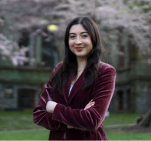 Photo of woman with long black hair standing with arms crossed smiling at the camera wearing burgundy velvet blazer. Background is outdoors on campus with flowering trees 