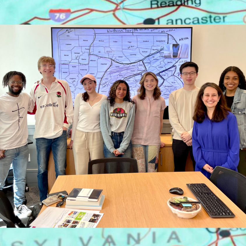 Research team of seven students with their professor standing at the back of meeting room smiling with a drawn map of Pennsylvania projected on a screen in the background.