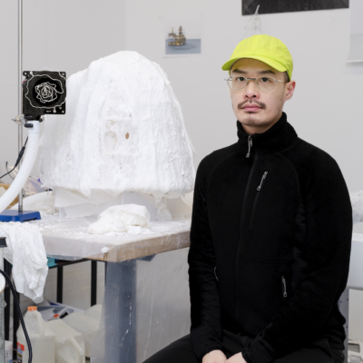 Portrait of male artist and masters student in his studio with a sculpture wearing all black with yellow baseball cap looking at camera