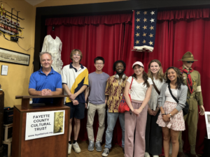 group of students with head of cultural trust posed for group photo surrounded by American artifacts that have been donated to the museum