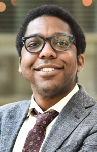 headshot for PIT UN speaker Bill Easley. Man with dark skin and black hair smiling at camera wearing black glasses and a grey blazer, brown tie, and white shirt