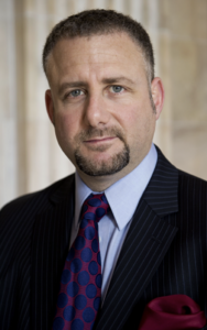 Headshot for man with short dark brown hair with beard and mustache wearing light blue shirt, red and blue tie, and dark blazer, standing against a backdrop with a column.