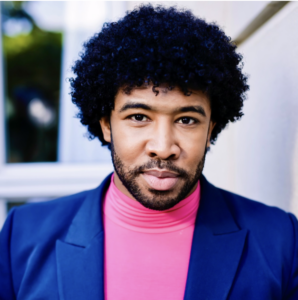 Male with dark skin and black short hair with beard and mustache wearing a pink sweater and royal blue blazer against a white backdrop and window with greenery to the left 