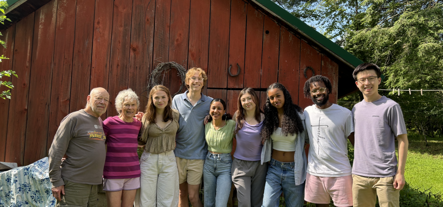 group photo of 9 people, in front of a red barn smiling and posed in a line for the camera