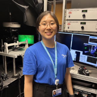 young woman smiling at camera in lab in front of computers and equipment.