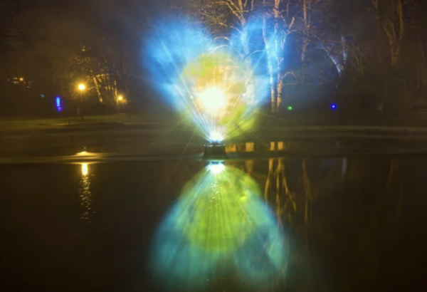 photo of a heart being projected outdoors lit up at night in colors of blue and yellow and a reflection of it on water