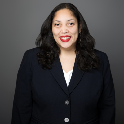 Headshot of woman with long black hair smiling at camera wearing a black blazer and white v-neck blouse standing against a grey backdrop