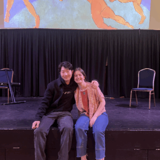 Two students seated on a stage smiling at the camera with a projection of a Matisse painting with dancers in a circle.
