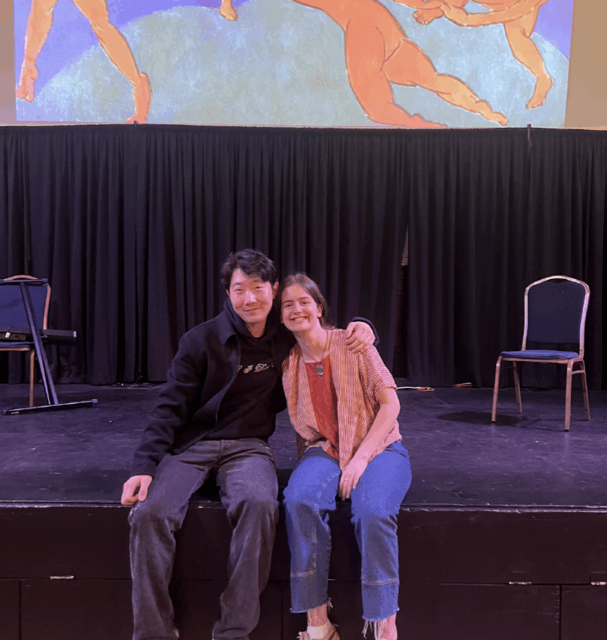 Two students seated on a stage smiling at the camera with a projection of a Matisse painting with dancers in a circle.