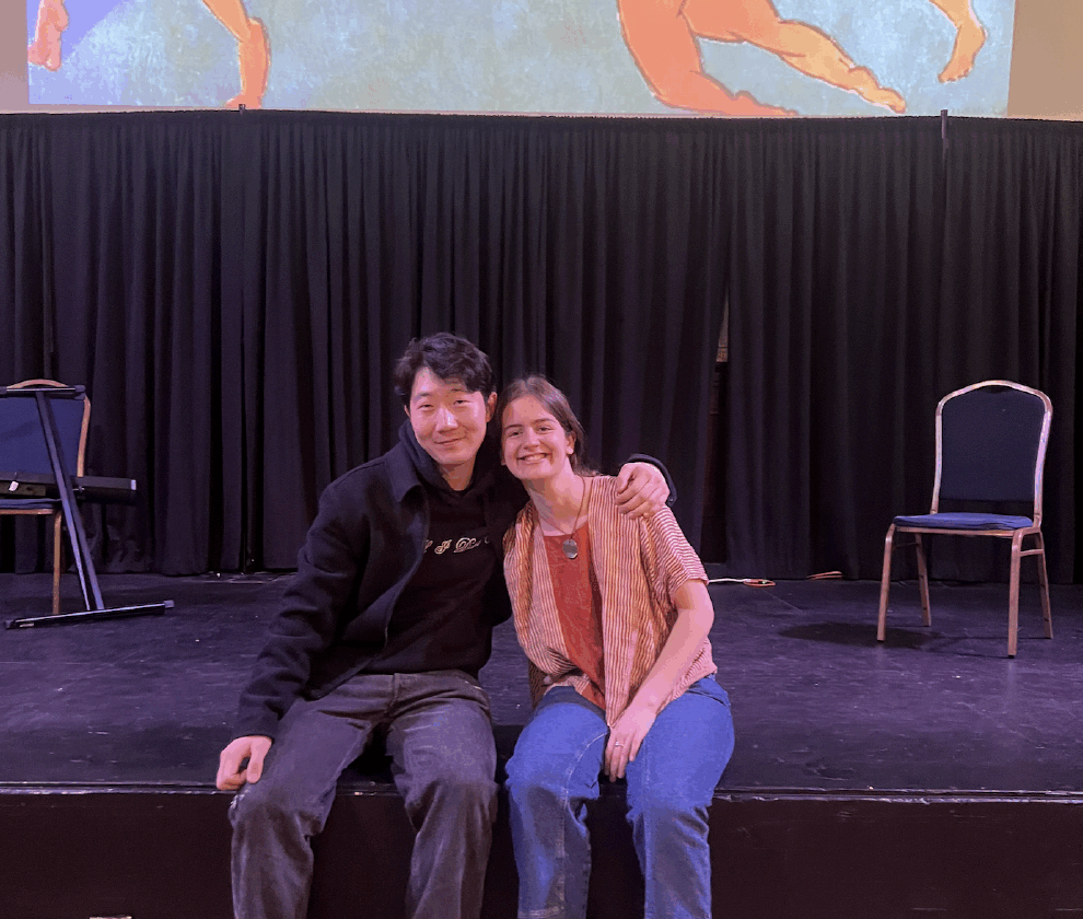 Two students seated on a stage smiling at the camera with a projection of a Matisse painting with dancers in a circle.