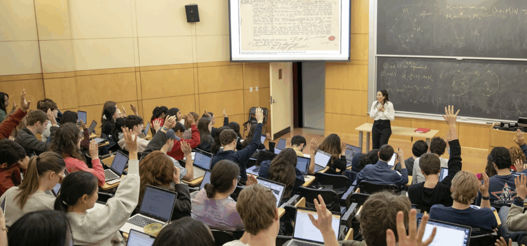 Professor at the head of a lecture size classroom with students raising their hands