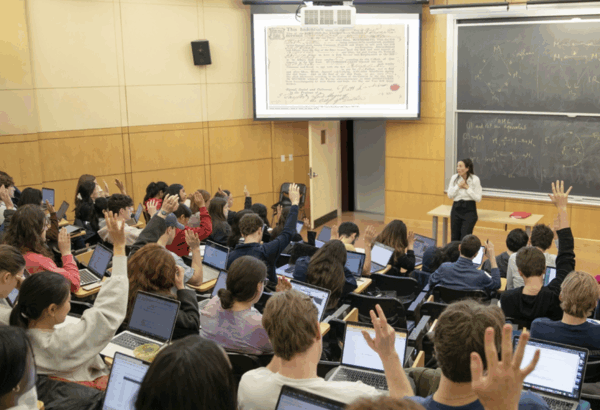 Professor at the head of a lecture size classroom with students raising their hands