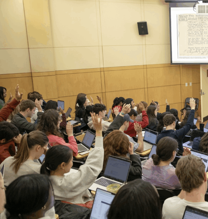 Professor at the head of a lecture size classroom with students raising their hands