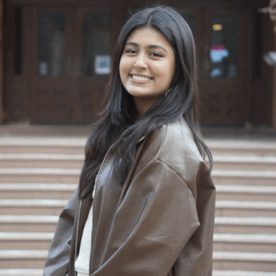 headshot of college student with long dark brown hair and light brown skin wearing a brown leather jacket standing outdoors in front of a building on campus